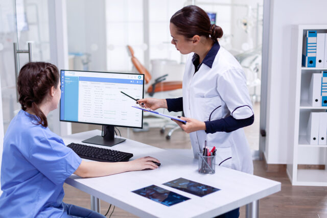 Two dental professionals reviewing patient records on a computer screen in a dental office. One is seated at the desk in scrubs using the computer mouse, while the other, wearing a white lab coat, stands beside her holding a clipboard and pointing at the monitor.