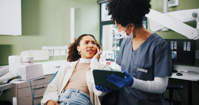 A patient sitting in a dental chair holding her cheek in discomfort while speaking with a dental professional. The dental professional, wearing scrubs, gloves, and a face mask, is holding a digital tablet and listening attentively