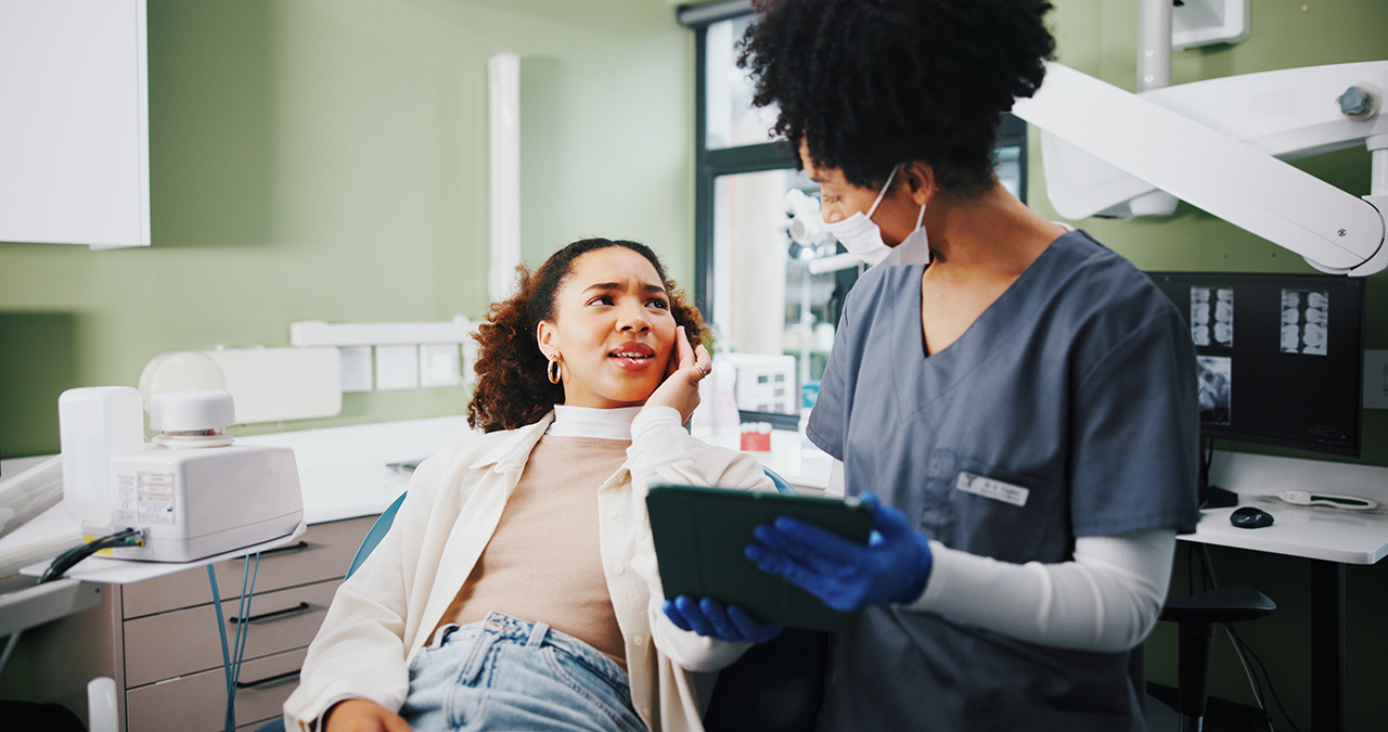 A patient sitting in a dental chair holding her cheek in discomfort while speaking with a dental professional. The dental professional, wearing scrubs, gloves, and a face mask, is holding a digital tablet and listening attentively