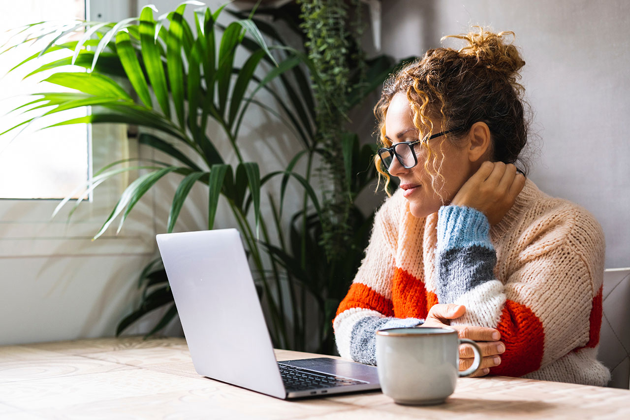 Woman wearing glasses and a colorful sweater working on a laptop at a wooden table with a coffee mug, sitting by a window and indoor plants in a cozy home office.