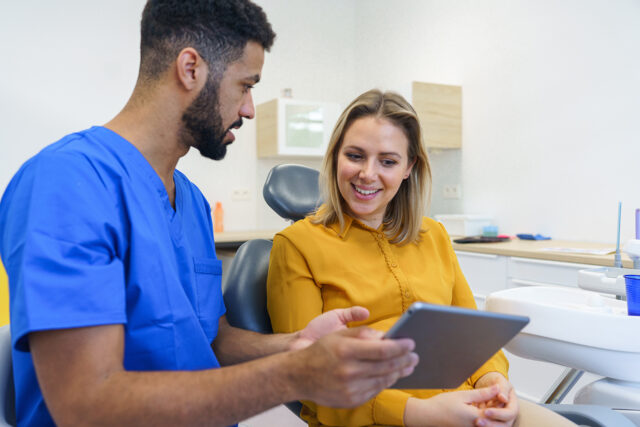 A dental professional in blue scrubs sits beside a patient in a treatment chair, showing her information on a digital tablet. The patient smiles while looking at the screen in a bright, modern dental office.