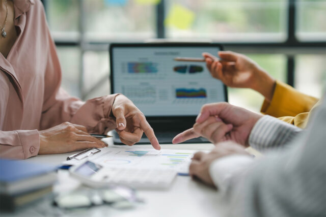 Business professionals collaborate around performance reports, pointing to data visualizations on printed charts and a laptop screen during a strategic metrics review meeting.