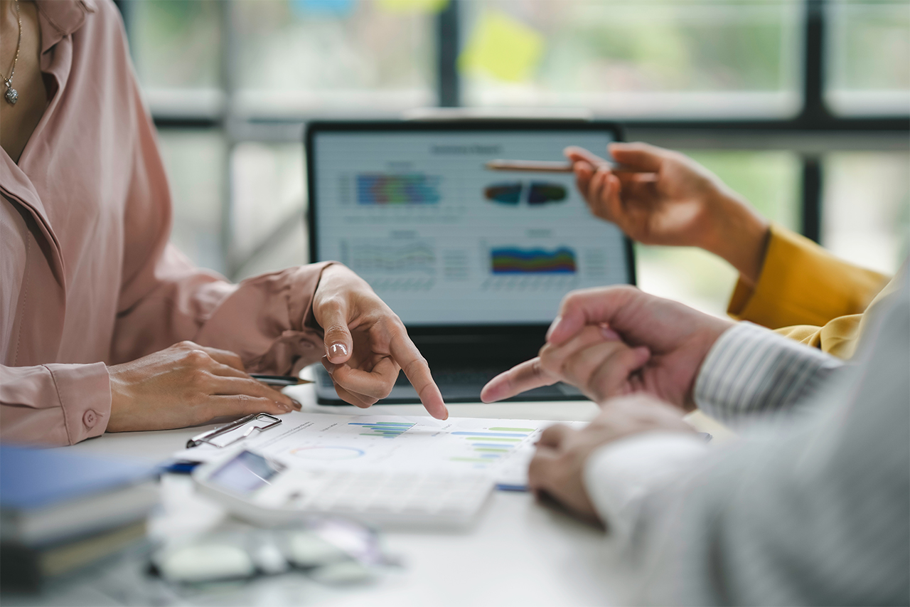 Business professionals collaborate around performance reports, pointing to data visualizations on printed charts and a laptop screen during a strategic metrics review meeting.