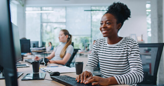 Smiling woman working at a computer in a modern DSO office with coworkers in the background.