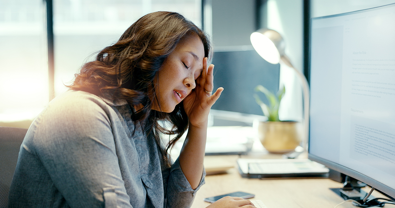 Healthcare administrative staff member experiencing burnout, sitting at a medical office desk with head in hand while working at a computer, conveying fatigue, stress, and emotional exhaustion.