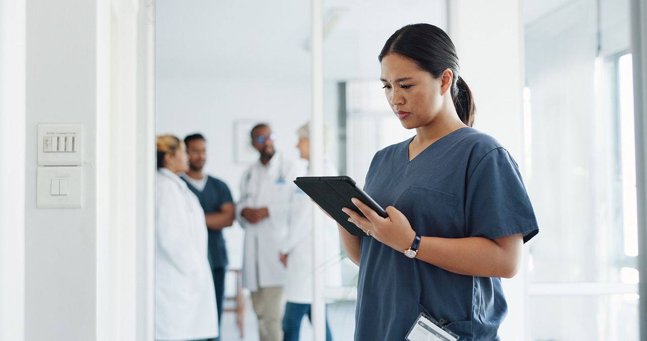 Healthcare manager reviewing information on a tablet in a clinical hallway, with medical staff collaborating in the background, representing coordination and management across multiple healthcare locations.