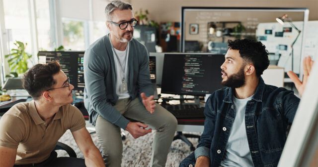 Three IT professionals collaborate in a modern office, reviewing code and discussing solutions on multiple computer screens, representing enterprise healthcare IT teams working on secure, scalable digital health systems.