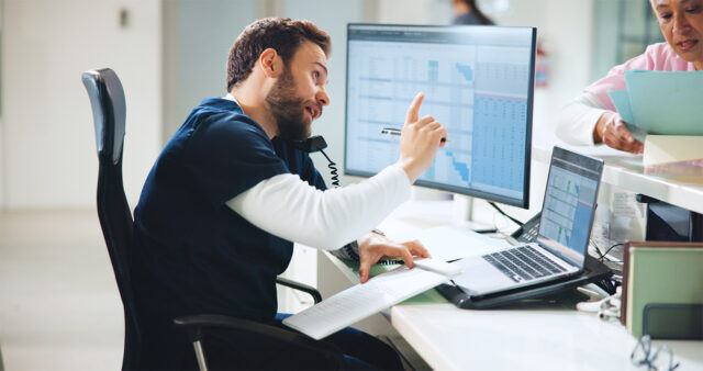 A healthcare worker in scrubs sits at a desk, speaking on a phone while pointing at a computer monitor displaying charts and data. A colleague stands nearby handing over a folder of documents in a clinical office setting.