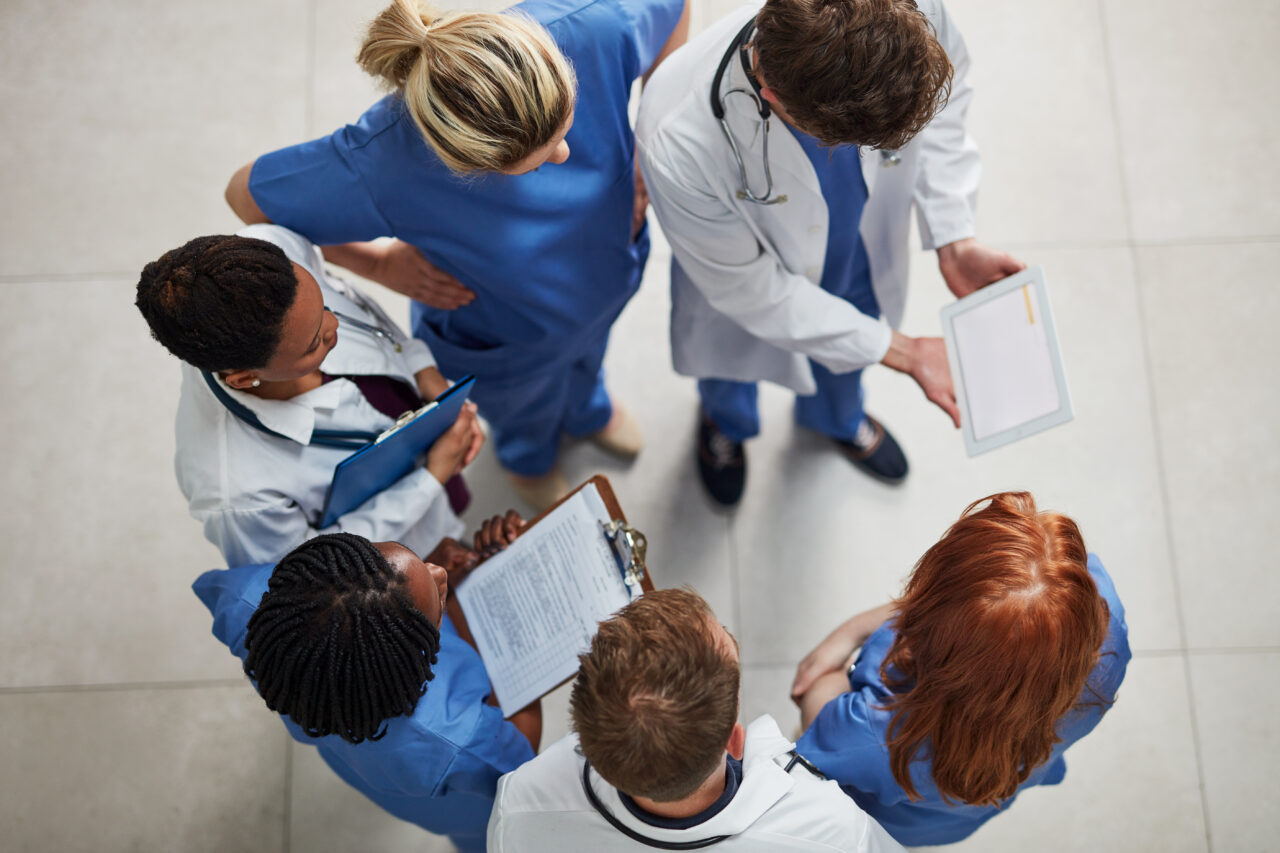 Overhead view of a diverse team of healthcare professionals in scrubs and lab coats gathered in a circle, reviewing a tablet and clipboards together—symbolizing collaboration and unified healthcare data sharing.