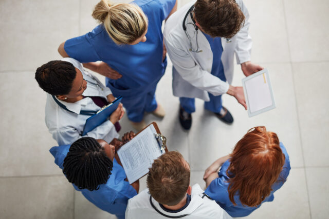 Overhead view of a diverse team of healthcare professionals in scrubs and lab coats gathered in a circle, reviewing a tablet and clipboards together—symbolizing collaboration and unified healthcare data sharing.