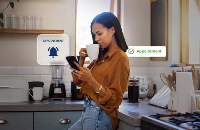 Woman standing in a kitchen drinking coffee while looking at her smartphone, with on-screen graphics showing an appointment reminder bell icon and a green checkmark confirming an appointment.