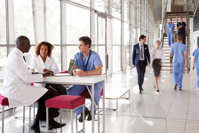 Healthcare professionals collaborate in a bright, modern hospital corridor, with two doctors and a nurse reviewing information on a laptop at a small table while other clinical staff move through the space—illustrating real-time communication and teamwork in a connected healthcare environment.