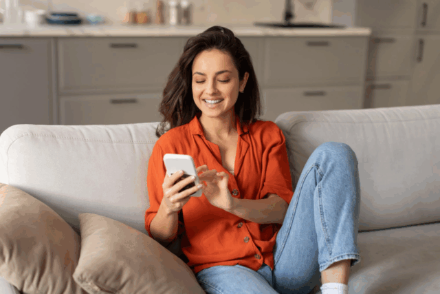 Woman sitting on a couch smiling while using her smartphone at home.