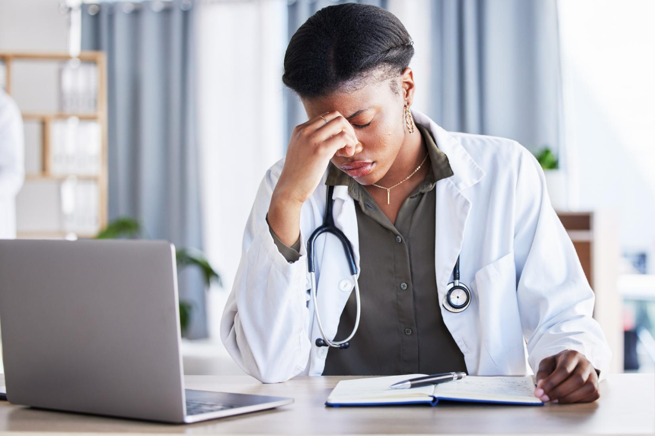 Doctor sitting at a desk with head in hand, appearing stressed while reviewing notes beside a laptop.