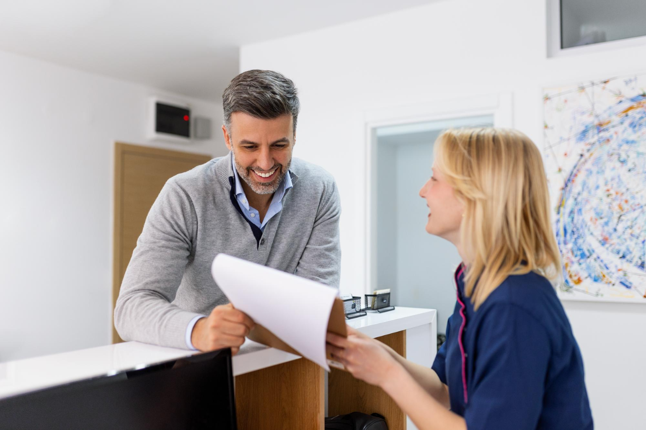 Healthcare staff member handing paperwork to a smiling patient at a medical office reception desk.